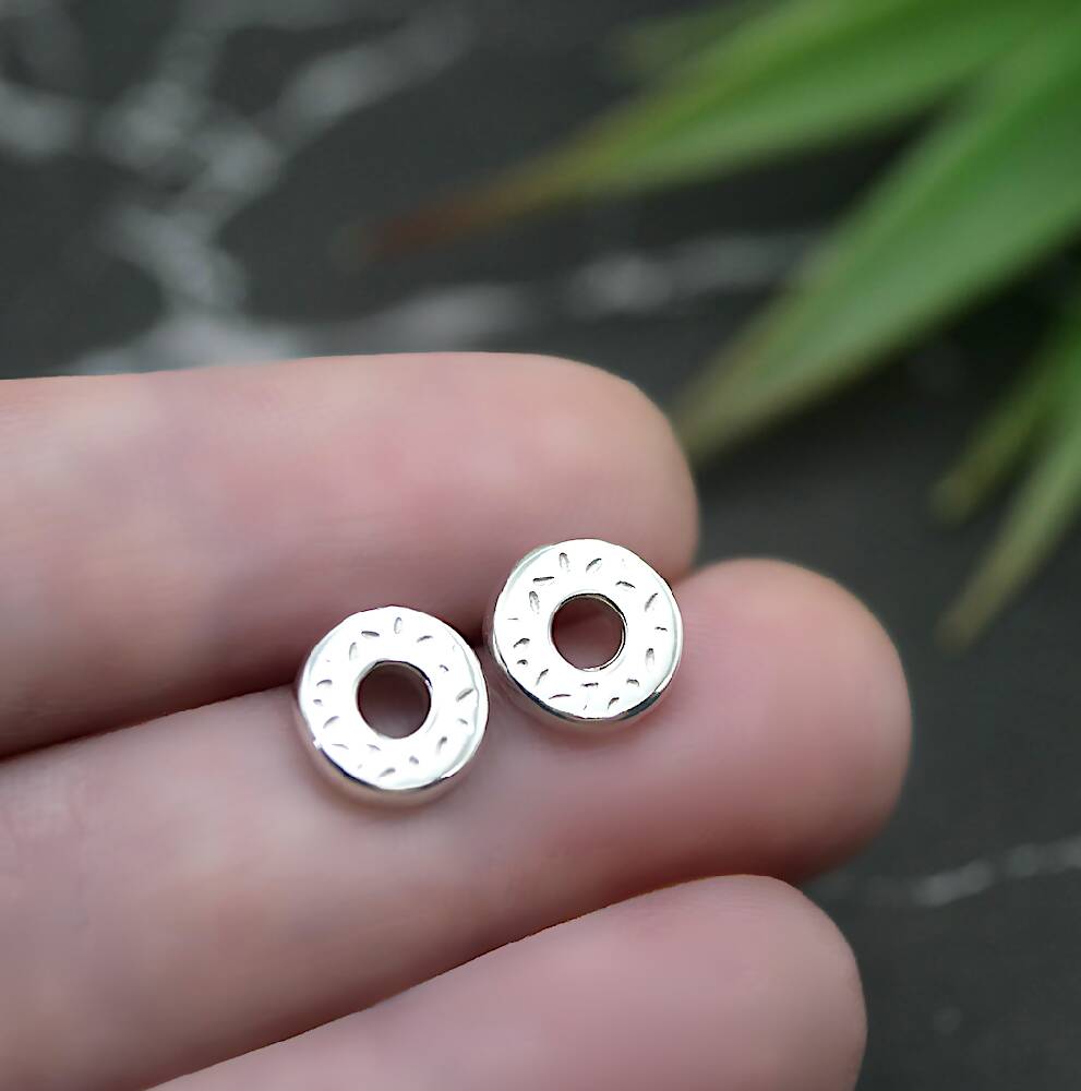 Image of handmade shiny sterling silver donut shaped studs by Purplefish Designs Jewellery. Earrings are held between fingers in an open hand, with a marble grey background with a decorative green plant.