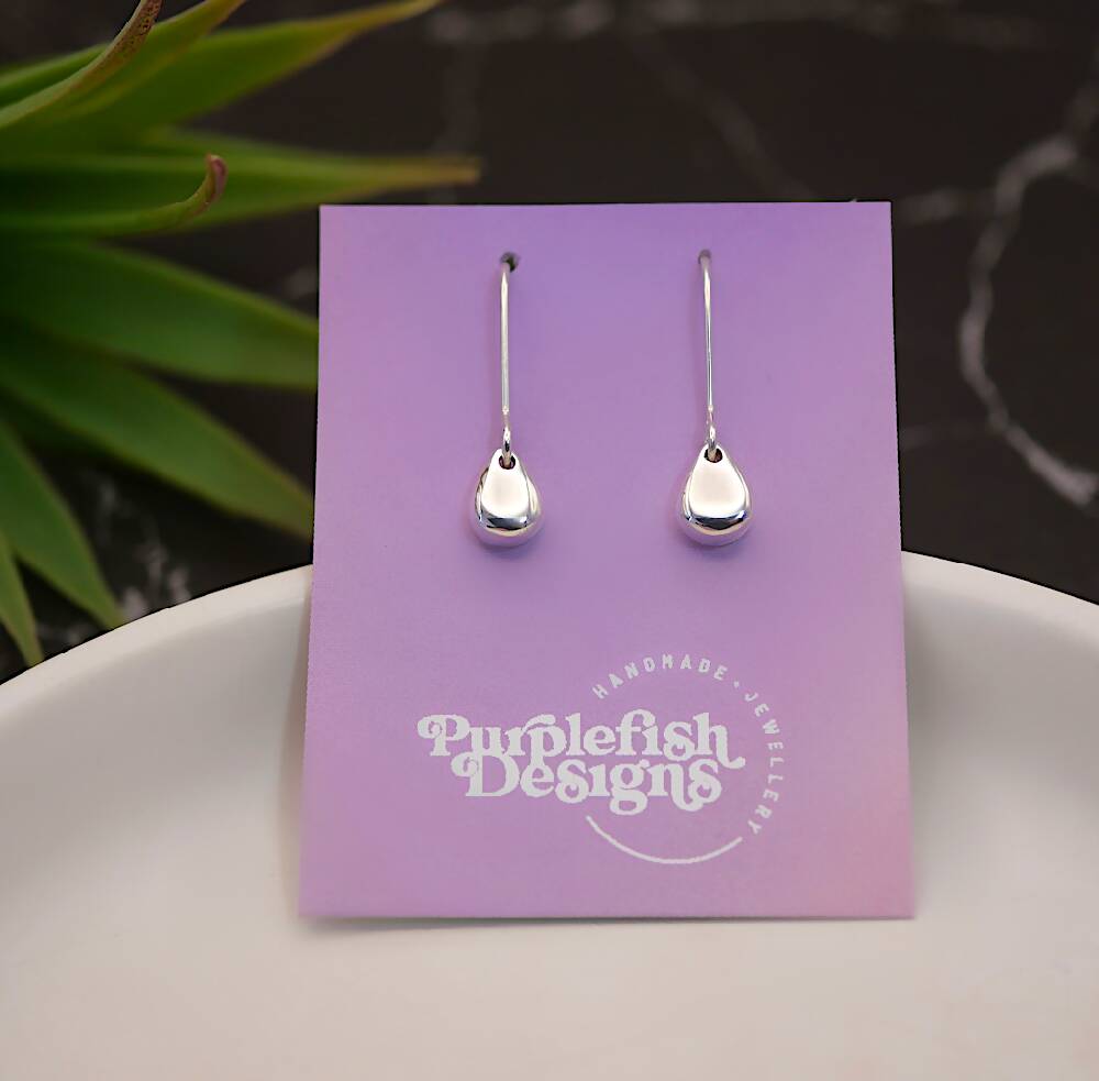 Image of a pair of sterling silver egg shaped earrings displayed on a purple and pink ombre earring card with a white Purplefish Designs logo. Earring card is resting in a white dish, on a marble grey background with a decorative green plant.