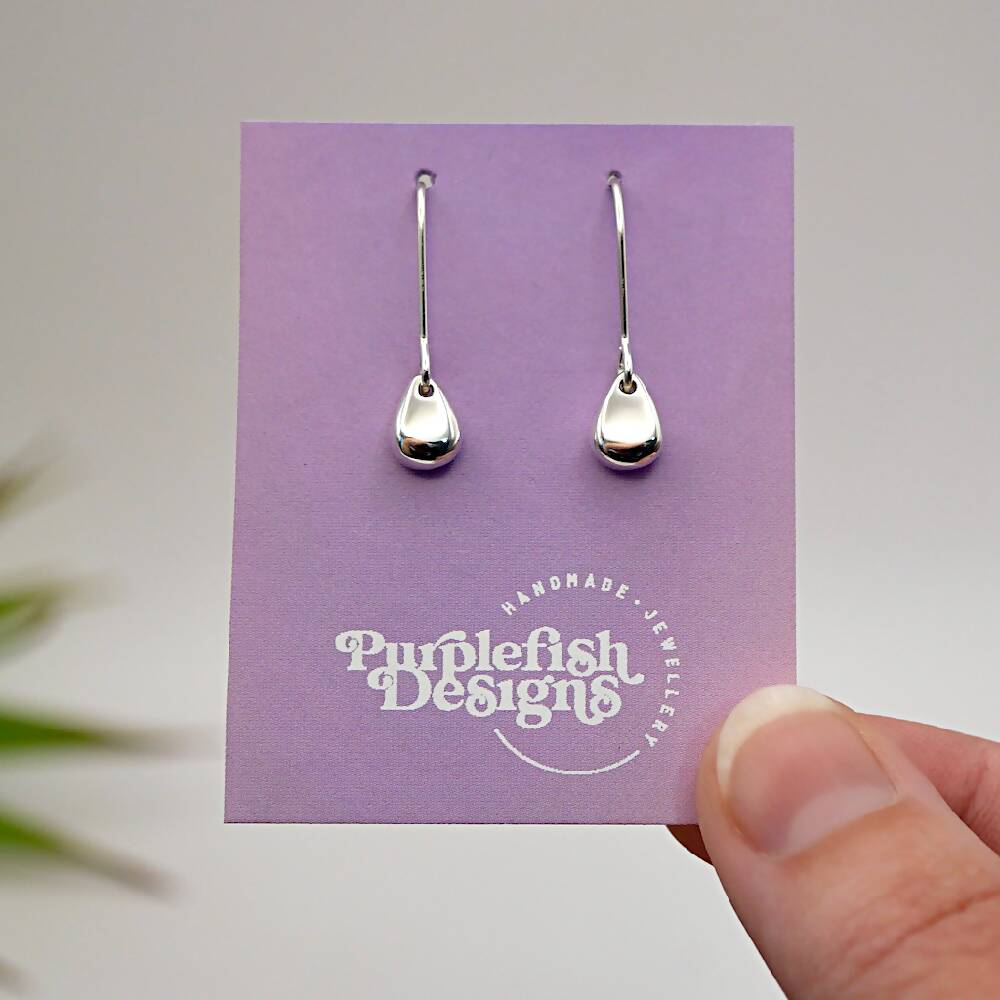 Image of a pair of sterling silver egg shaped earrings displayed on a purple and pink ombre earring card with a white Purplefish Designs logo. Earring card is held between thumb and finger in front of a white background with a decorative green plant.