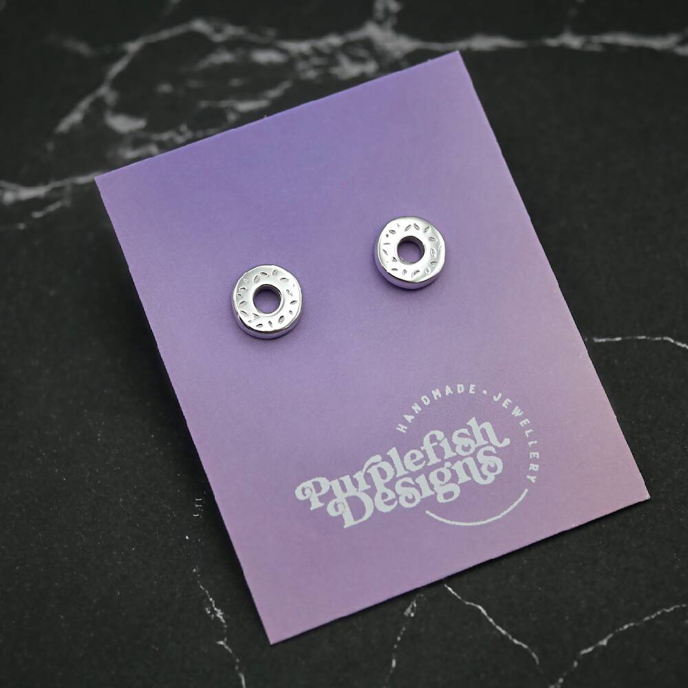 Image of a pair of sterling silver donut shaped earrings displayed on a purple and pink ombre earring card with a white Purplefish Designs logo. Earring card is resting on a marble grey background with a decorative green plant.