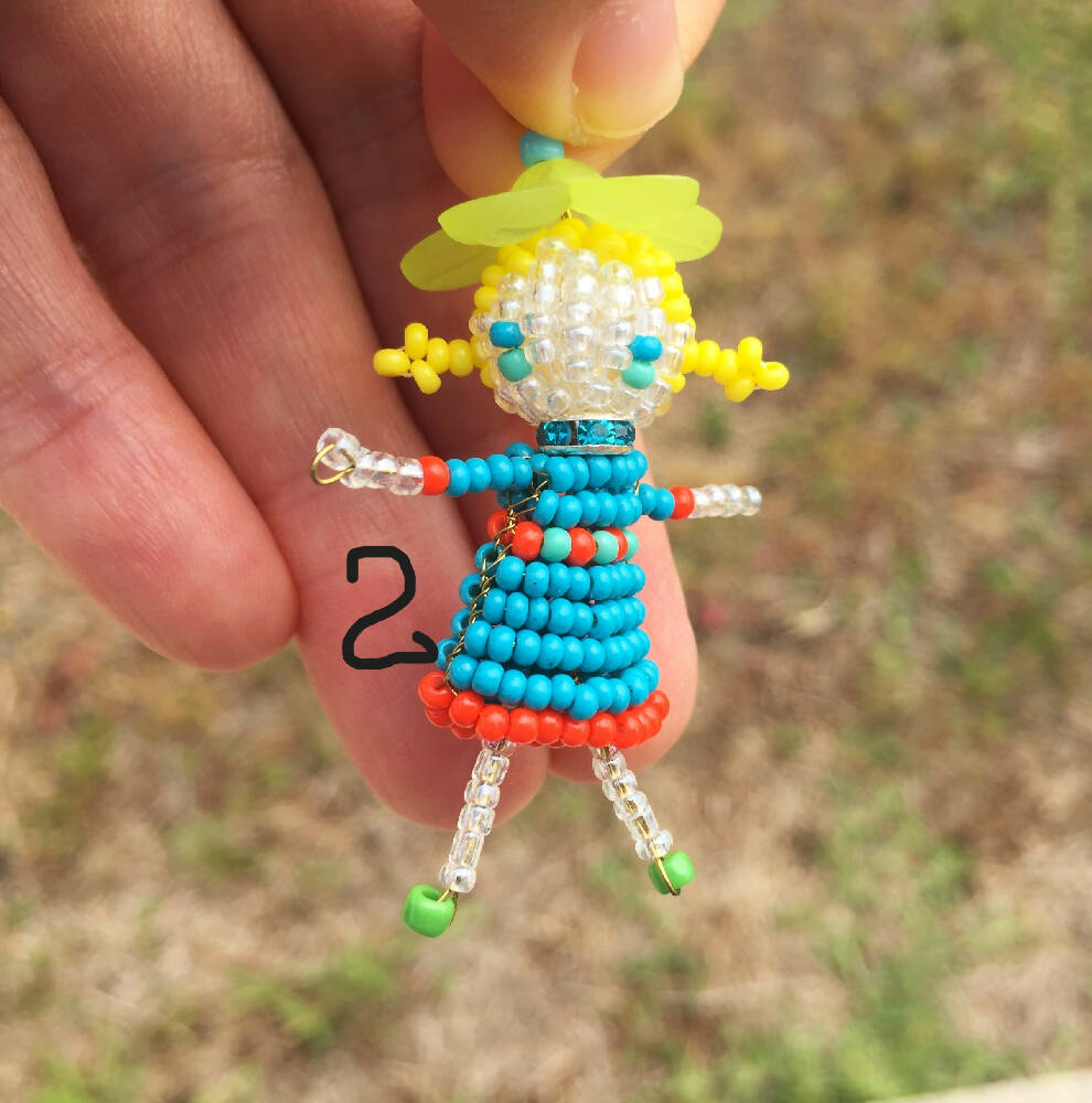Naryanabeads beaded doll option2 - beaded doll with light blue crystal collar, light green flower bead hat, yellow braided hair and blue/turquoise eyes. Legs, arms, face made of shiny clear beads, blue dress with orange trim, shoes - green beads..