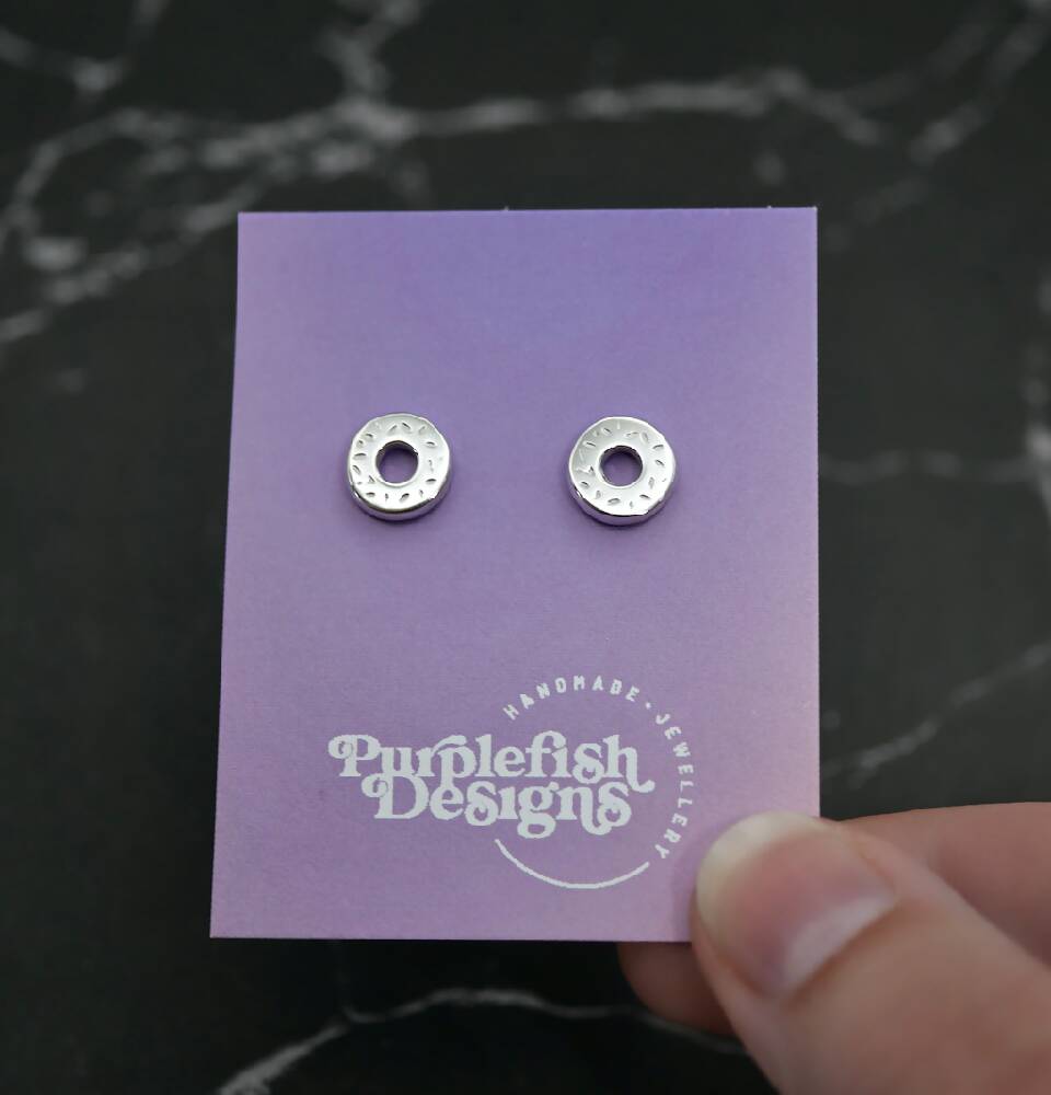 Image of a pair of sterling silver donut shaped studs displayed on a purple and pink ombre earring card with a white Purplefish Designs logo. Earring card is held between thumb and finger in front of a marble grey background with a decorative green plant.