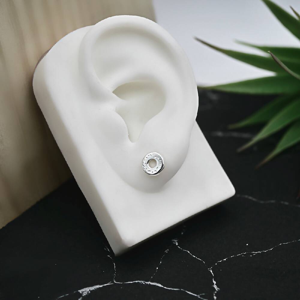 Image of shiny sterling silver donut shaped studs by Purplefish Designs Jewellery. Earring is displayed on a white silicone ear to illustrate how the stud will sit on an earlobe. Ear model is leaning against a wooden board, on a marble grey background with a decorative green plant.