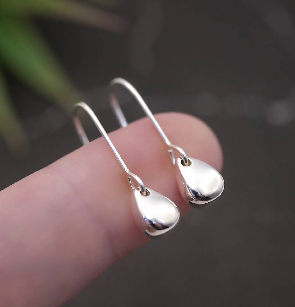 Image of handmade shiny sterling silver egg shaped earrings by Purplefish Designs Jewellery. Earrings are resting on one finger to give an indication of size and scale and are on a grey marble background with decorative green plant.