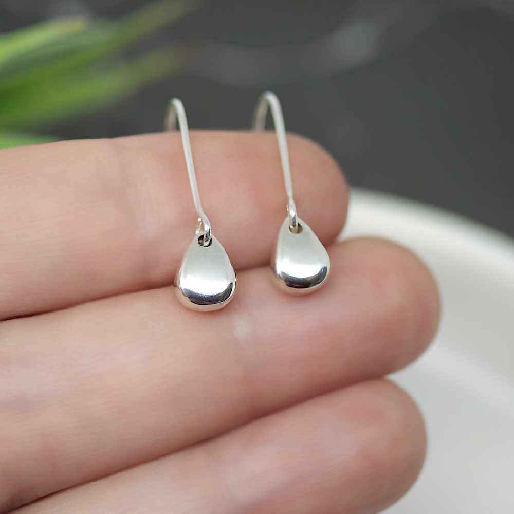 Image of handmade shiny sterling silver egg shaped earrings by Purplefish Designs Jewellery. Earrings are resting on the fingers of an open hand to give an indication of size and scale and are on a grey marble background with decorative green plant