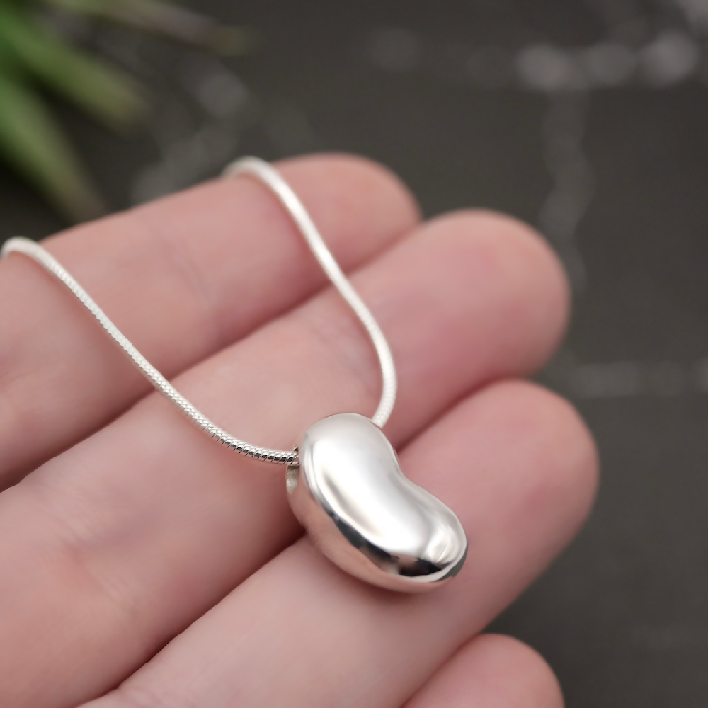 Image of handmade shiny sterling silver Bean shaped pendant by Purplefish Designs Jewellery on silver snake chain necklace. The pendant is draped over an open hand to give an indication of size and scale, on a grey marble background with decorative plant.