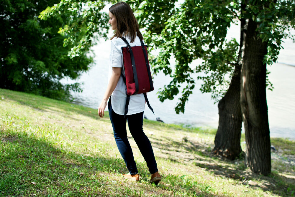 woman is walking on a riverside with her bordeaux colour backpack with black leather straps. she had brown long hair and wearing a white shirt.