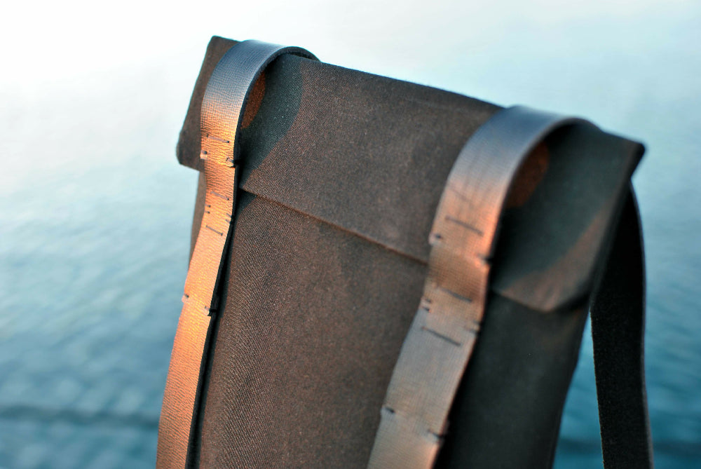 the upper part of a black canvas backpack with black leather straps. water in the background.