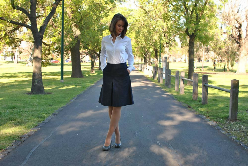 A woman is wearing navy bue denim coulotte skirt, white shirt and gray high heel shoes while she is standing on a road in a park. Her left hand is in her pocket.
