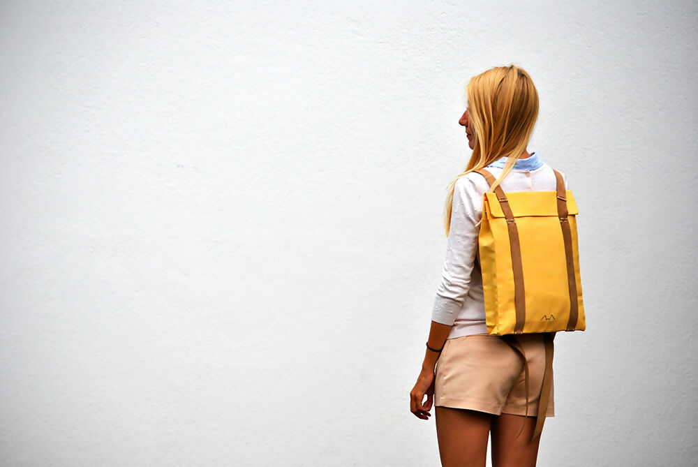 A bonde girl is wearing a yellow canvas backpack with suede leathr straps while she is fcing a white wall. She wears a white jumper and beige shorts.