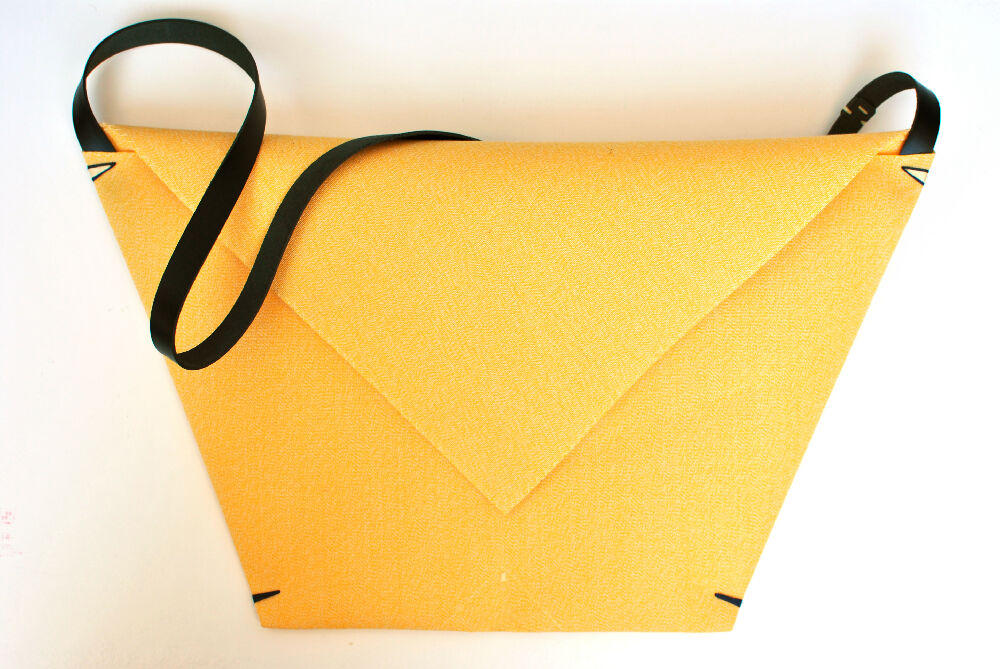 Yellow minimalist envelope bag with black leather strap is standing on a white table.