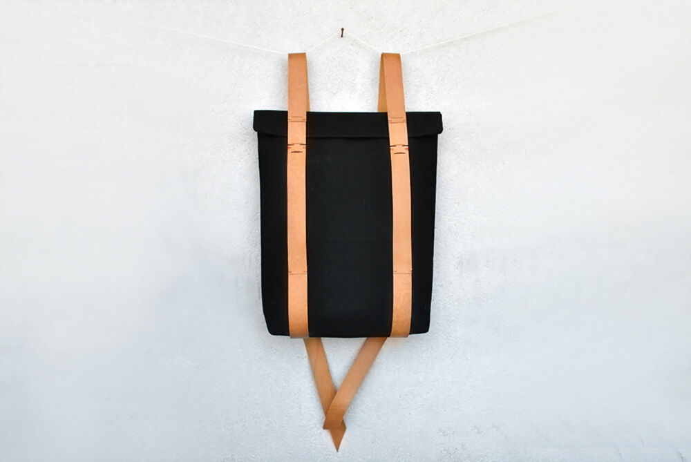 A black minimalist backpack with nude colour leather straps is hanging in front of a white wall.