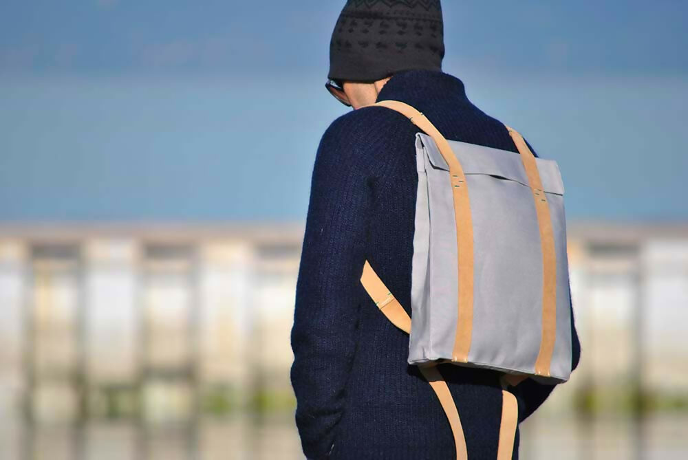 A man in dark beanie is standing on a pier and watchin down. He wears a navy blue jumper and a grey backpack with suede leaher straps.