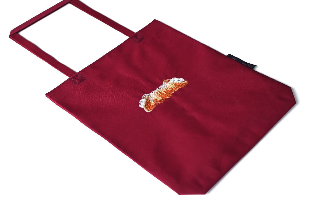 Bordeaux paperbag lookalike canvas tote with cannoli embroidery, lying on a white table.