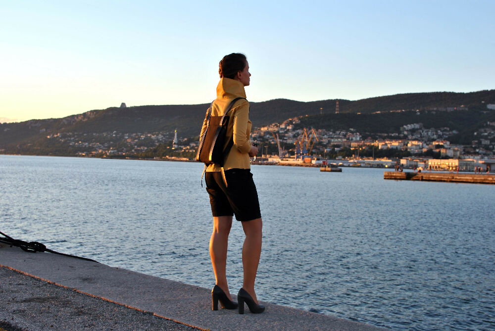 woman in a yellow blouse anad black shorts is standing in front of the sea on a pier and wearing a black minimalist backpack