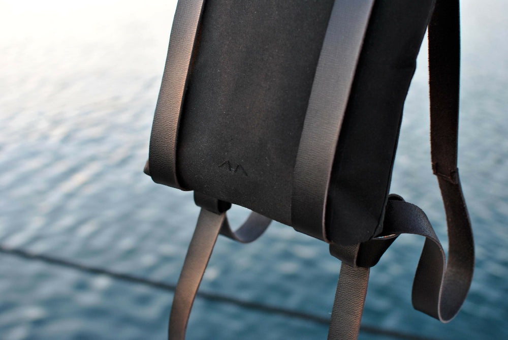 the lower part of a black canvas backpack with black leather straps. water in the background.