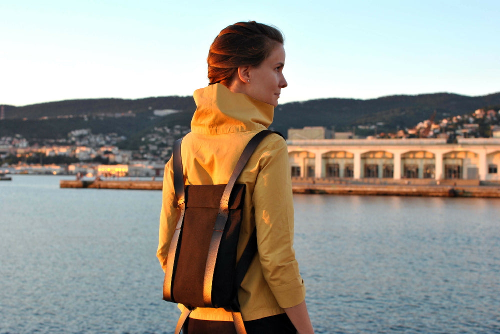 Woman in a yellow blouse is looking over her right shoulder. She is standing in front of the sea and wearing a black minimalist backpack