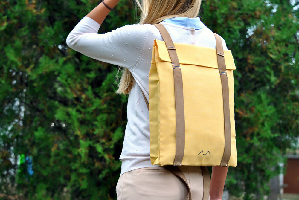 A bonde girl is wearing a yellow canvas backpack with suede leathr straps She wears a white jumper and beige shorts. there are cypress trees in the background. She runs her left hand through her hair