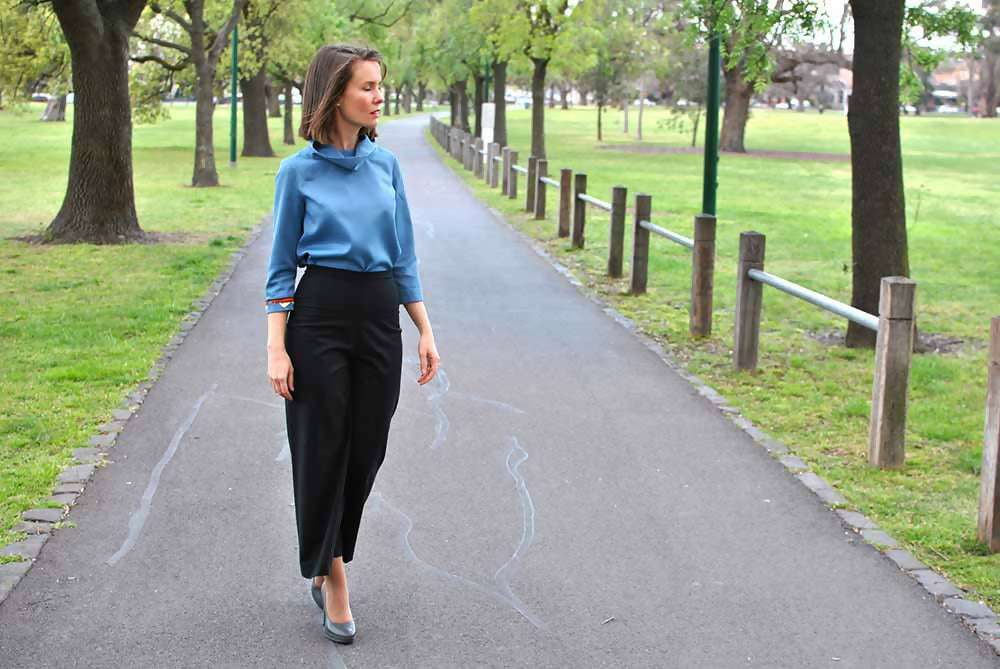 Woman in black wool crop pants and blue blouse is walking on a road in a park.
