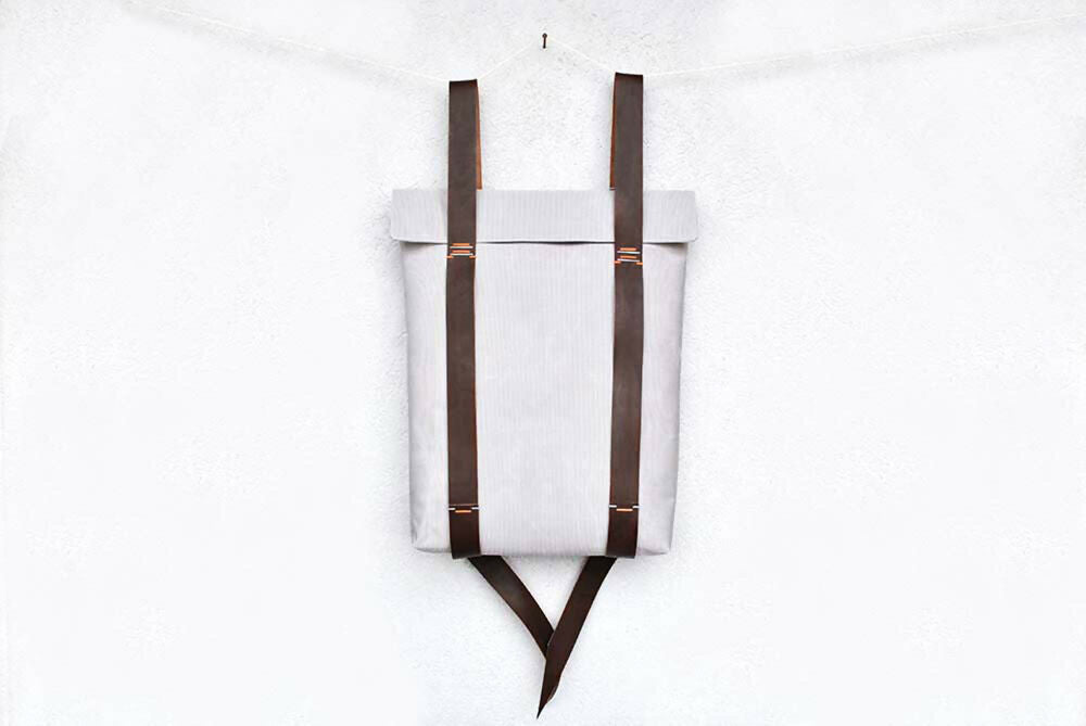 A light grey minimalist backpack with brown leather straps is hanging in front of a white wall.