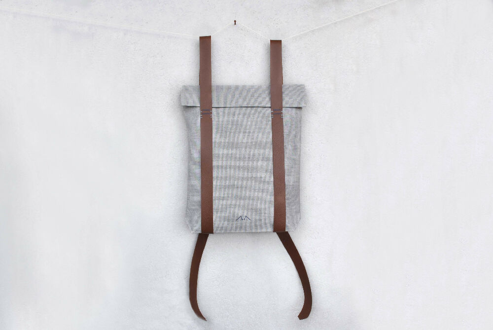 A grey minimalist backpack with brown leather straps is hanging in front of a white wall.