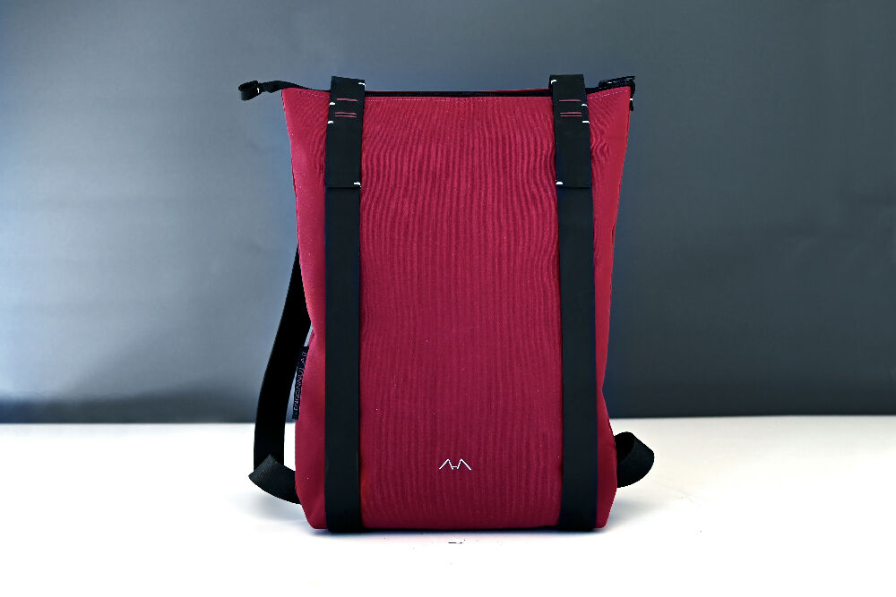 bordeaux colour backpack with black minimalist leather straps is standing on a white table in front of a gray background.