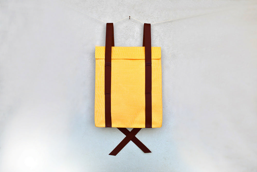 A ellow backpack with dark brown leather straps is hanging on a wall.