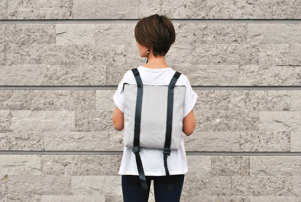 woman with short brown hair is wearing white T-shirt and gray canvas backpack with black leather straps. She is standing in front of a gray brick wall.