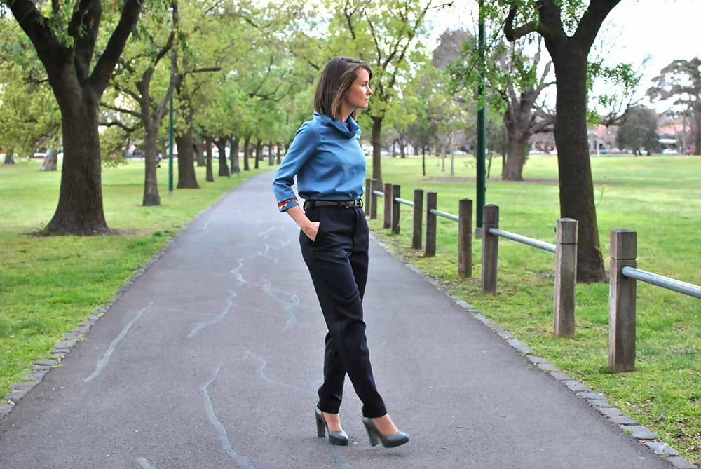 A woman is wearing black business pants, blue blouse and gray high heel shoes while she is standing on a road with her hands in her pokets.