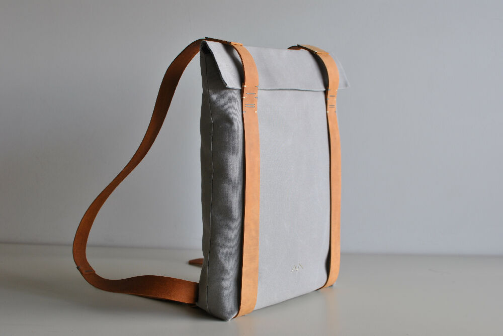 Grey minimalist backpack with nude leather straps is standing on a beige table in front of a white wall.