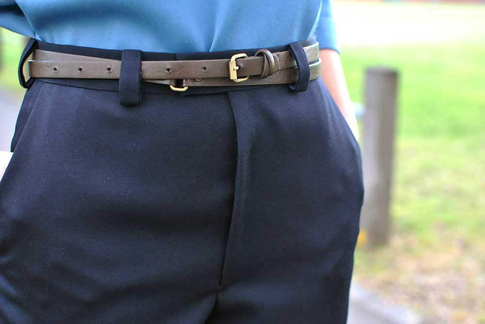 Details of a black formal pants. A woman is wearing them with blue blouse and brown belt while holding her hands in the pockets.