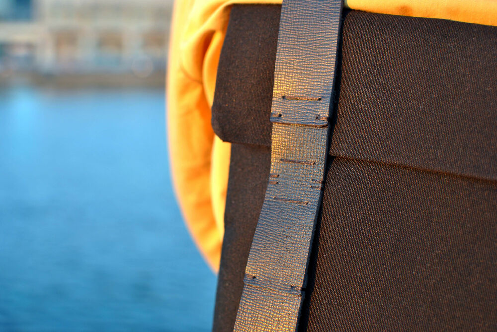 Details of a black backpack with handstitched black leather straps. On a person who is wearing a yellow blouse. There is water in the background.