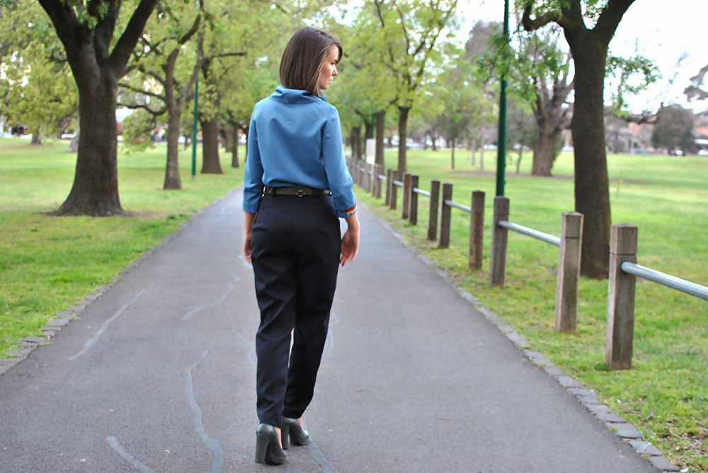 We can see the back of a woman in black wool formal pants and blue blouse is walking on a road in a park.