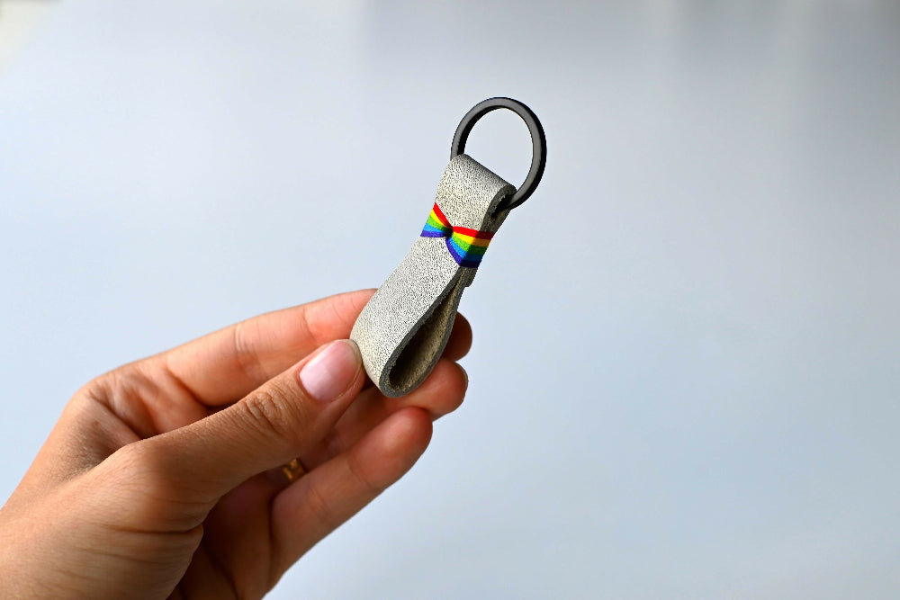A hand is holding a white leather keychain with colourful rainbow stitches and black split ring. White background.