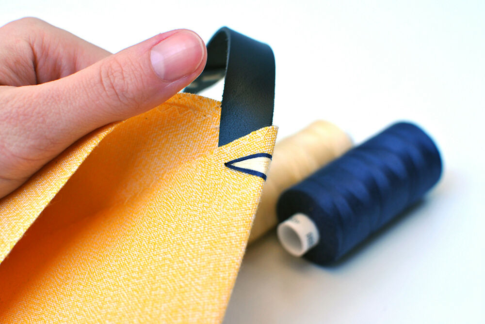 Hand is holding a yellow bag with black strap to show its handstitched blue and yellow details. There are yellow and dark blue threads in the background.