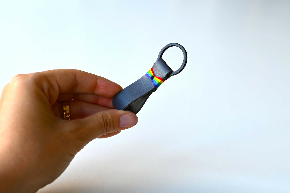 A hand is holding a grey leather keychain with colourful rainbow stitches and black split ring. White background.