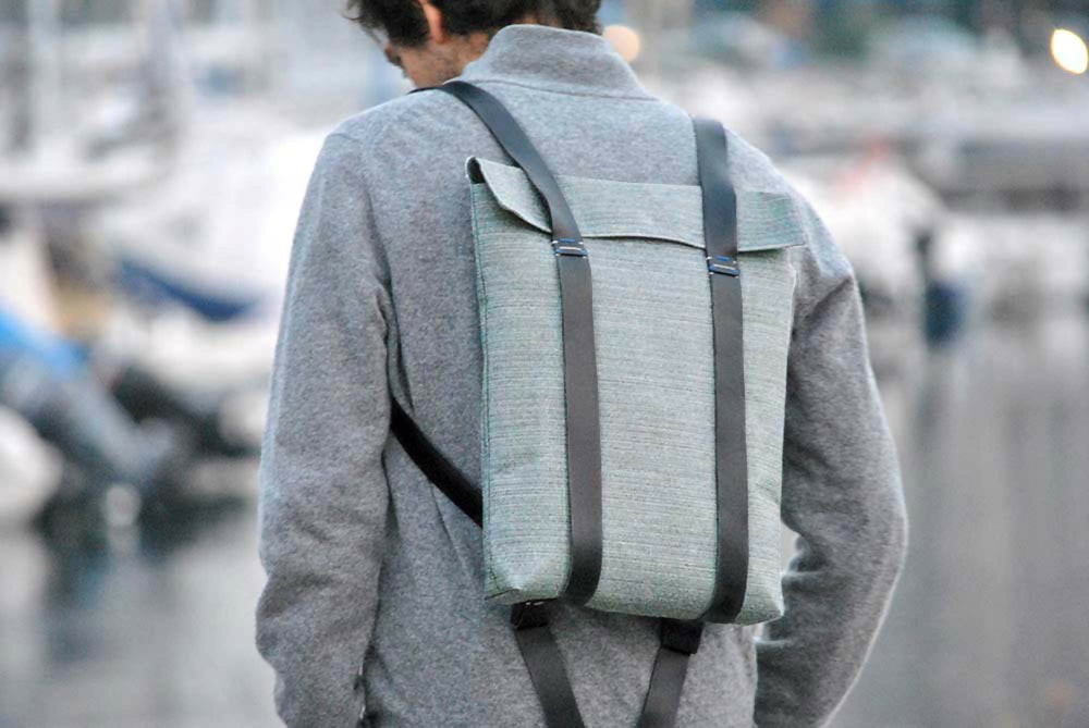 Man in gray jumper is wearing a gray canvas minimalist backpack with black leather straps. The background is blurred.