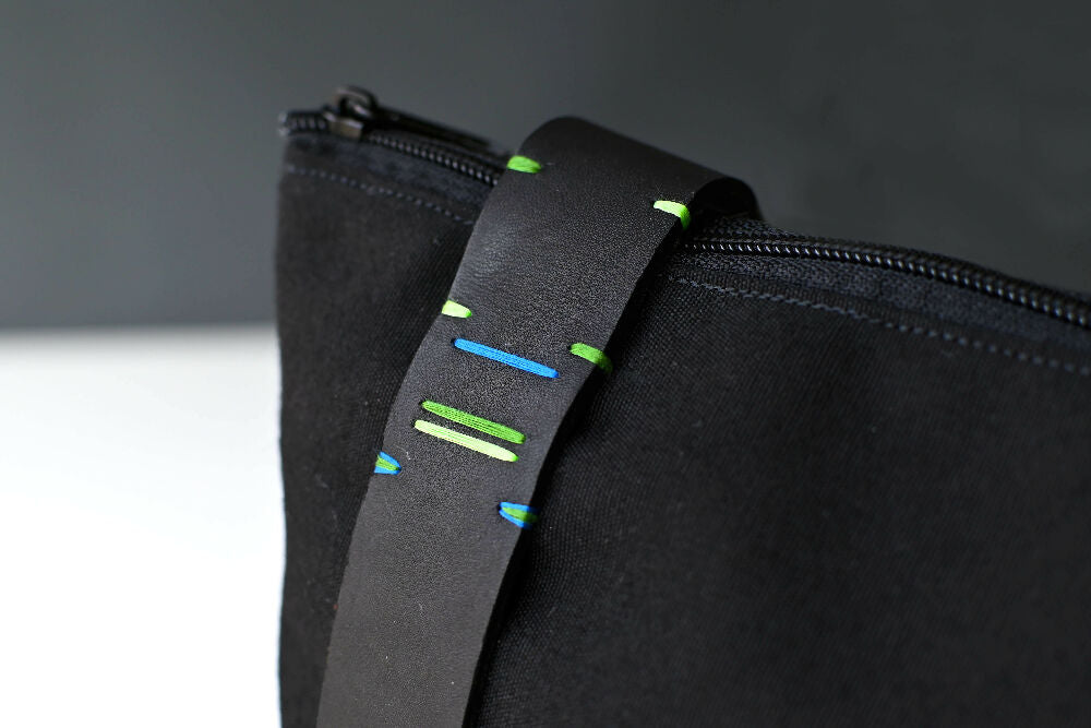 Colourful green and blue handstitched details on a black leather strap of a black canvas backpack.