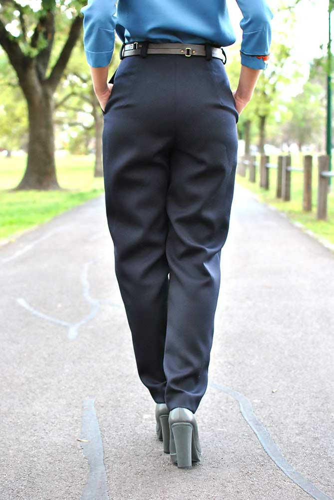 The lower part of the back of a woman who is wearing black formal pants, blue blouse and gray high heel shoes. Her hands are in her pants' pockets.
