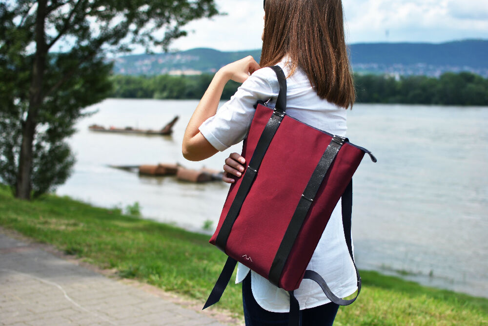 woman is pulling her bordeaux colour backpack with black leather straps forward. she had brown long hair and is standing on the riverside in white shirt.