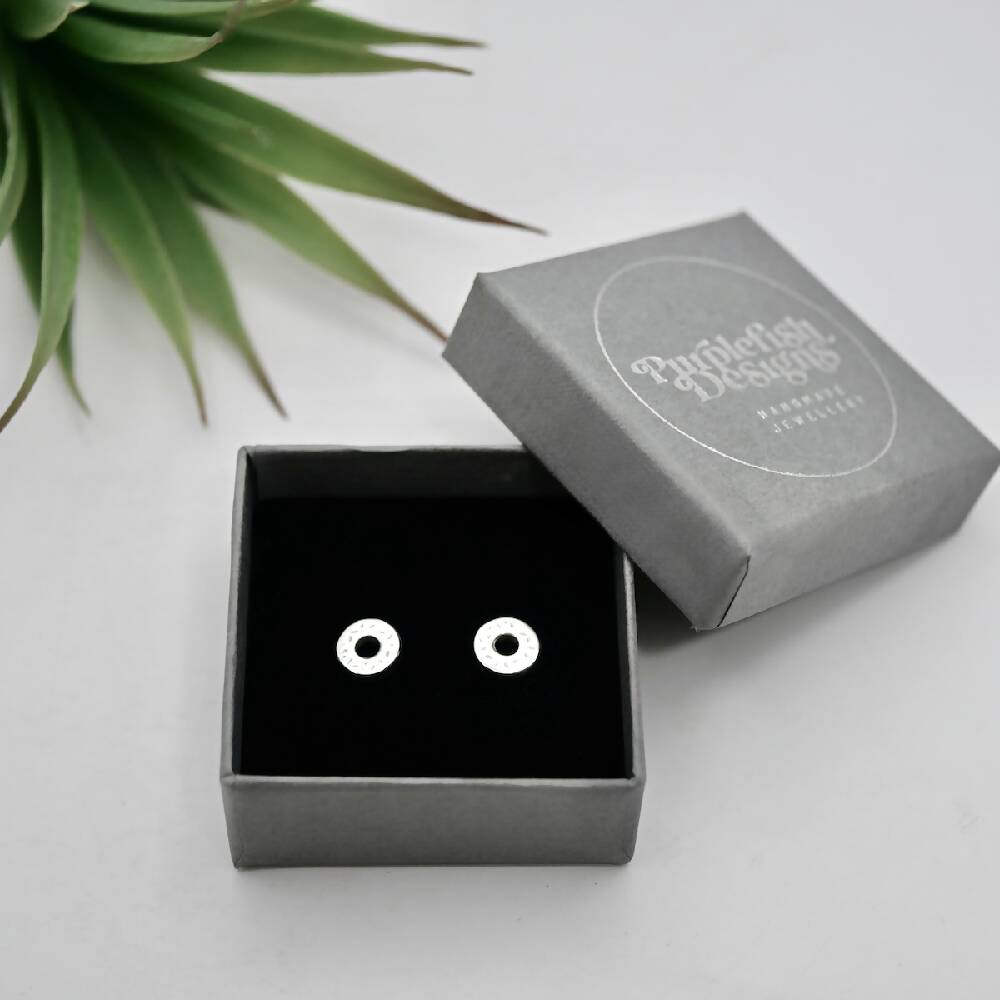 Image of a pair of handmade sterling silver donut shaped studs in a small grey jewellery gift box embossed with silver Purplefish Designs Handmade Jewellery logo. Box is on a white background with a decorative green plant.