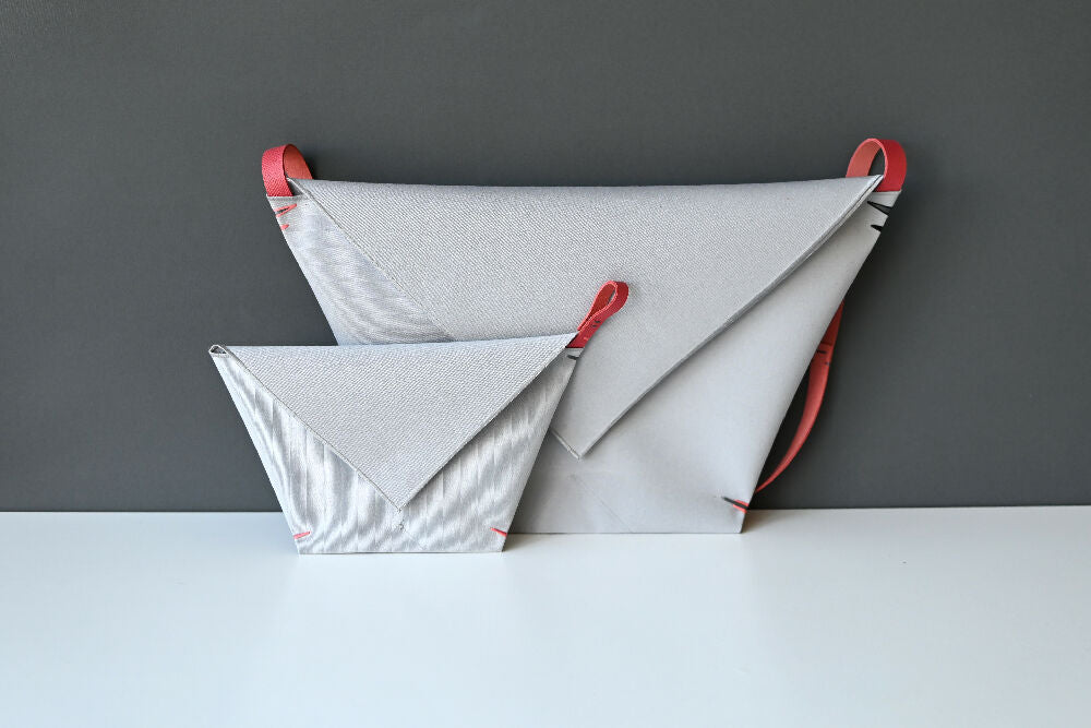 Gray minimalist envelope bag with matching detechable matching pochette, made from canvas and salmon colour leather strap are standing on a white table, in front of a gray background.