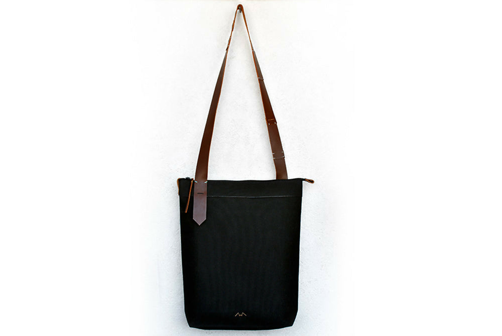 A black canvas bag with long asymmetric brown leather strap is hanging on a white wall.