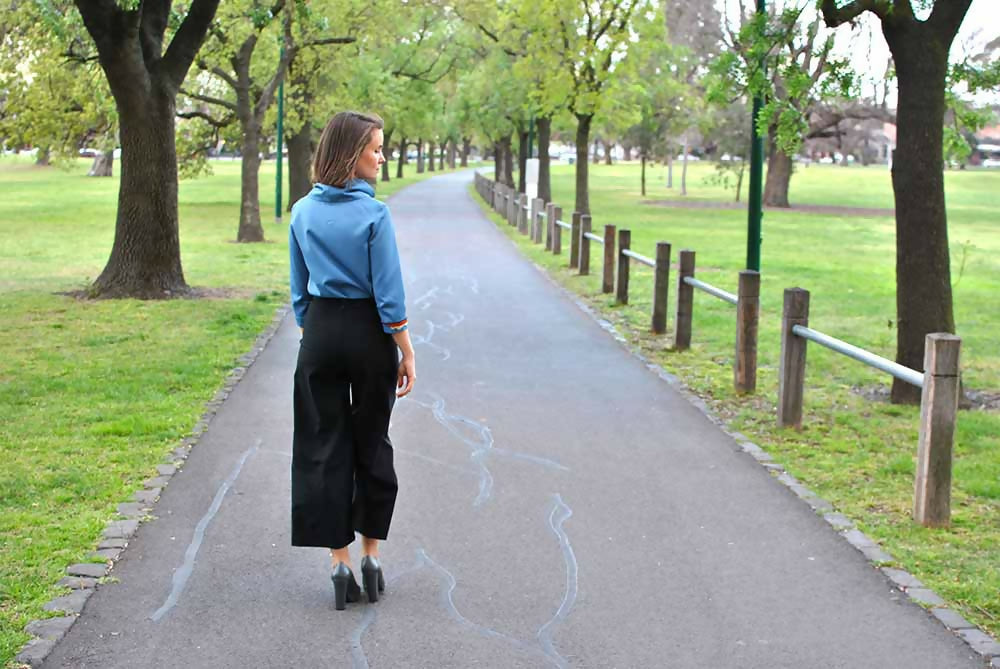 We can see the back of a woman in black wool crop pants and blue blouse is walking on a road in a park.