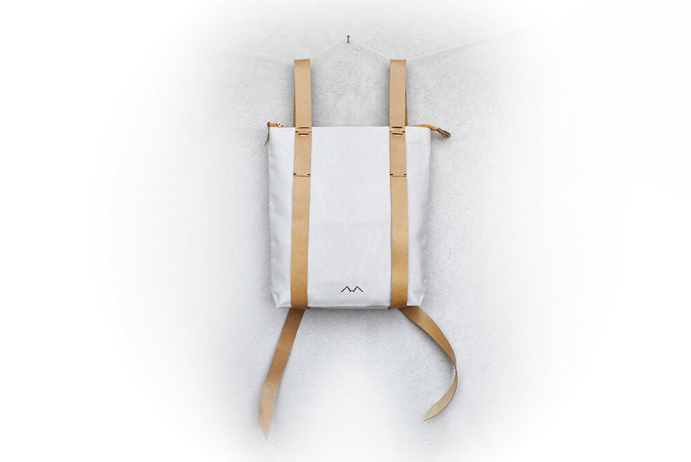 A gray backpack with nude colour leather straps is hanging in front of a white wall