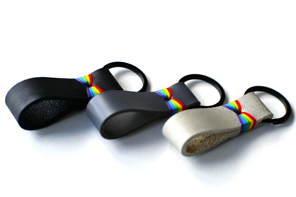 A black, a gray and a white leather keychain with colourful rainbow stitches are lying on a white surface. They have black split ring.