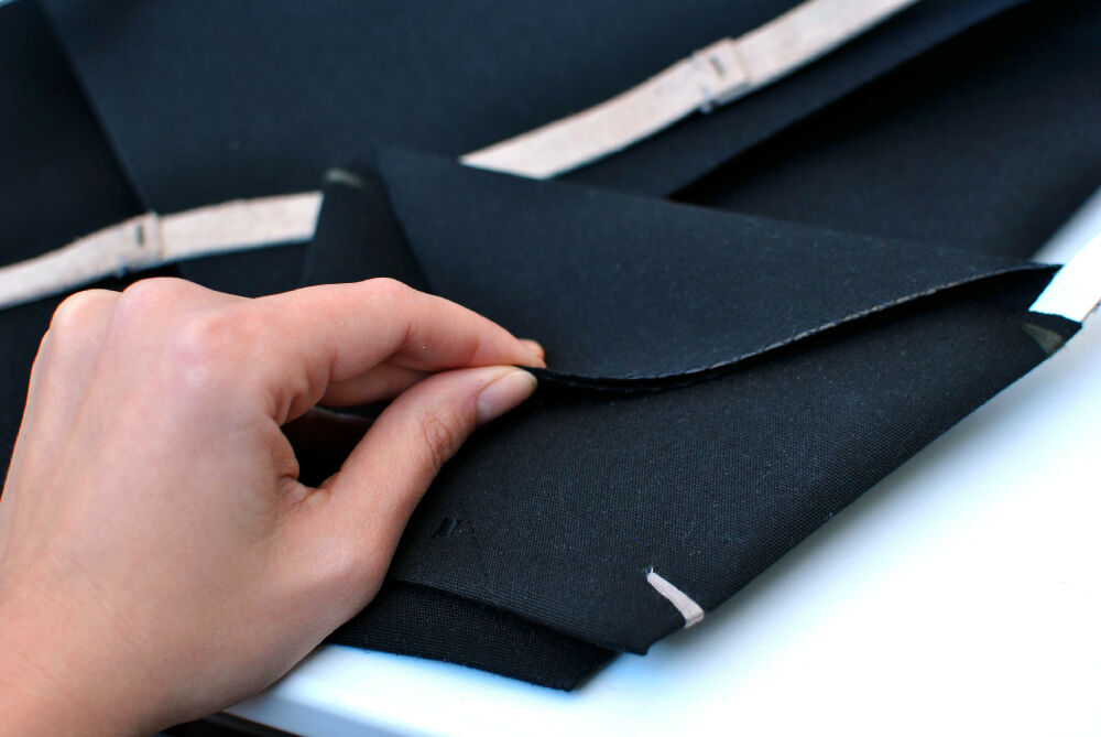 Hand is opening a black minimalist black canvas pochette  which is lying on a black matching envelope clutch.