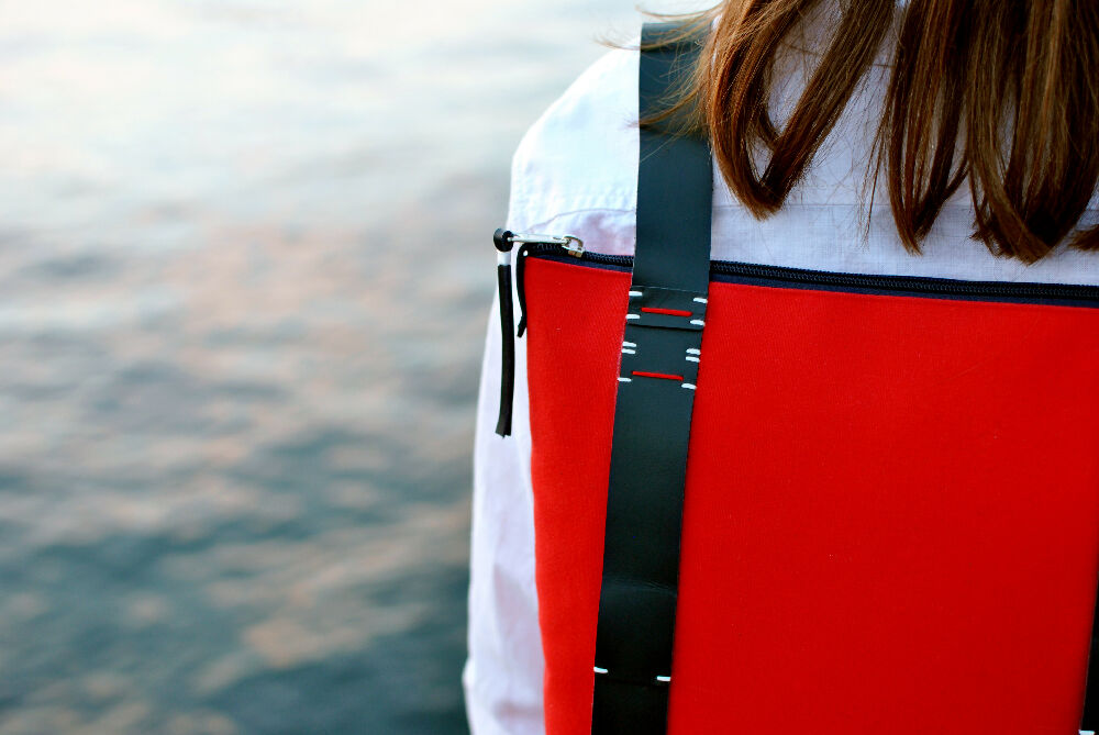 shoulder of a woman who is wearing white shirt and red backpack with handstitched black leather straps. the backpack has zipper. water in the background