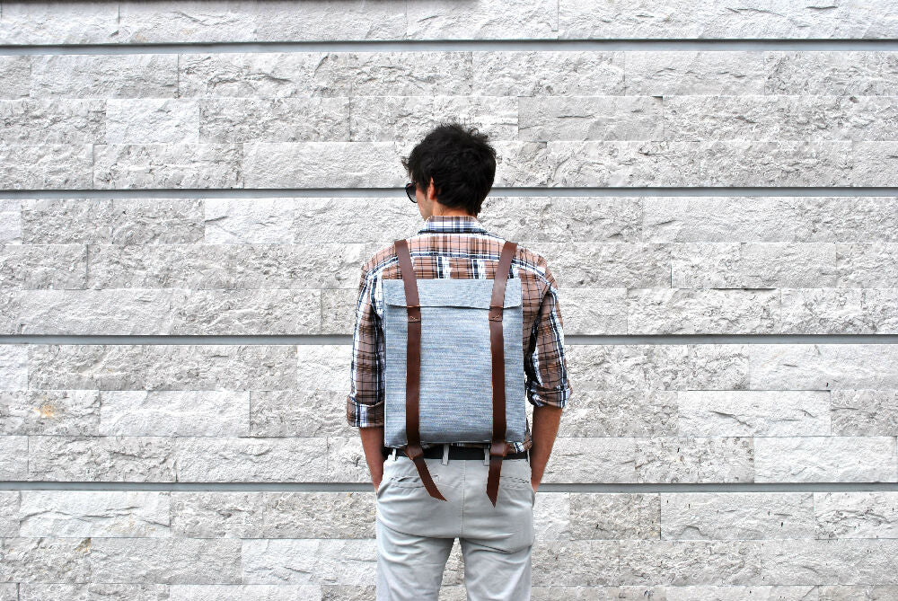 A man with dark brown curly hair is facing a gray brick wall while wearing a grey canvas bag with brown leather straps. He wears a checkered brown shirt with beige pants and his hands are in his pockets.