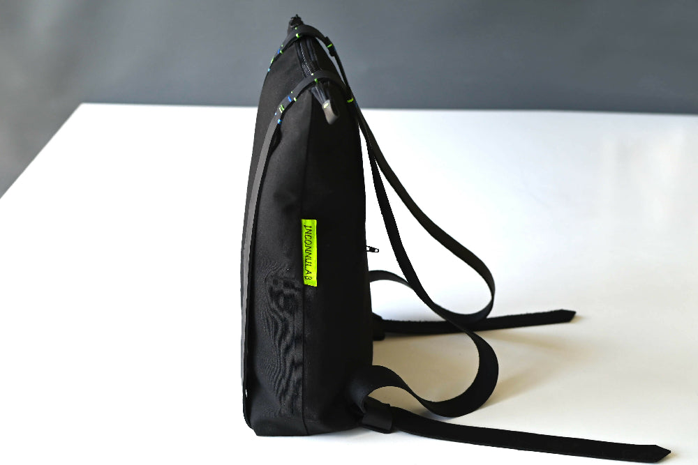 One side of a black backpack with black leather straps which has a green tag standing on a white table in front of a gray background